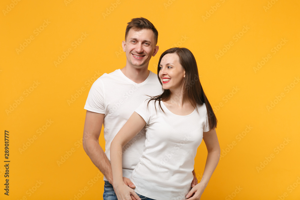 Beautiful young couple two friends guy girl in white empty blank design t-shirts posing isolated on yellow orange background. People lifestyle concept. Mock up copy space. Looking on camera and aside.
