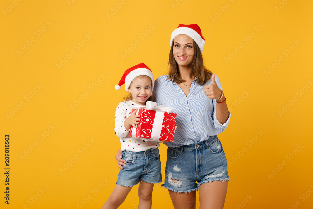 Obraz premium Woman child baby girl in Christmas Santa hat hold gift box. Mommy little kid daughter isolated on yellow background studio portrait. Happy New Year 2020 celebration holiday concept. Mock up copy space