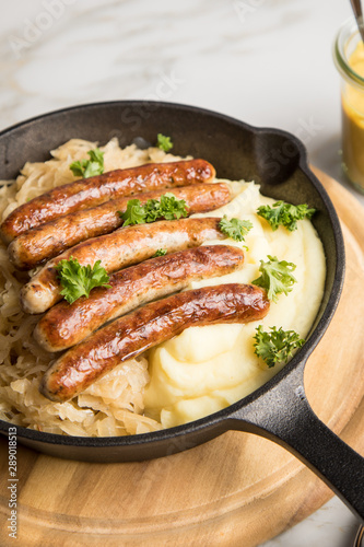 Kleine Bayrische Nürnberger Rostbratwürste mit Sauerkraut, Kartoffelpüree und Senf in rustikaler Gusseisenpfanne auf Holz Brett und Marmor Tisch