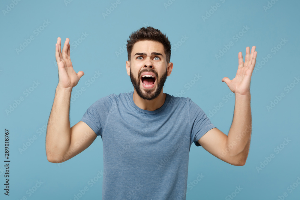 Naklejka premium Young angry shocked man in casual clothes posing isolated on blue background, studio portrait. People sincere emotions lifestyle concept. Mock up copy space. Looking up, screaming, spreading hands.