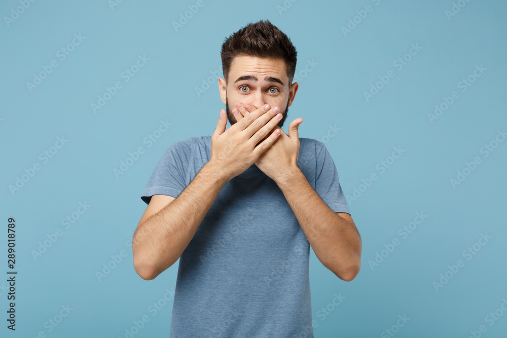 Fototapeta premium Young shocked concerned man in casual clothes posing isolated on blue wall background, studio portrait. People sincere emotions lifestyle concept. Mock up copy space. Covering mouth with hands.