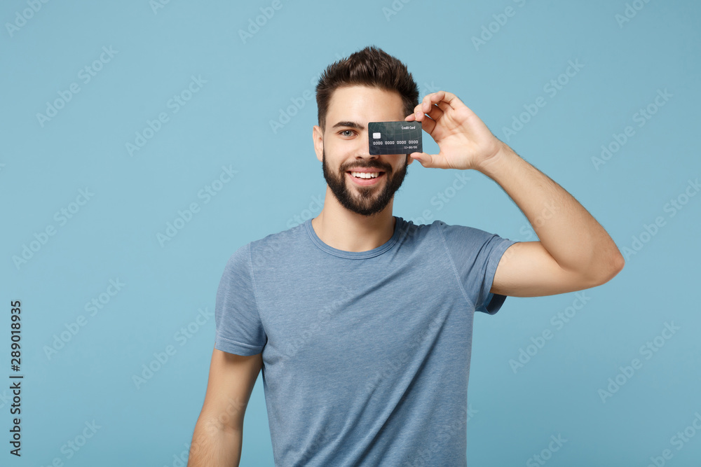 Young smiling handsome man in casual clothes posing isolated on blue wall background studio portrait. People sincere emotions lifestyle concept. Mock up copy space. Covering eye with credit bank card. Young smiling handsome man in casual clothes posing isolated on blue wall background studio portrait. People sincere emotions lifestyle concept. Mock up copy space. Covering eye with credit bank card.