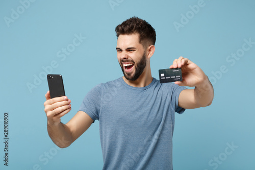 Young screaming man in casual clothes posing isolated on blue wall background, studio portrait. People sincere emotions lifestyle concept. Mock up copy space. Holding mobile phone, credit bank card. © ViDi Studio