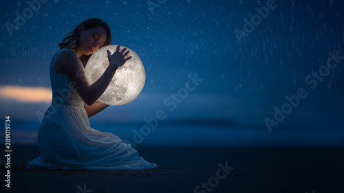 Tender image of a girl; female magic. Beautiful attractive girl on a night beach with sand and stars hugs the moon, art photo. On a dark background with space.