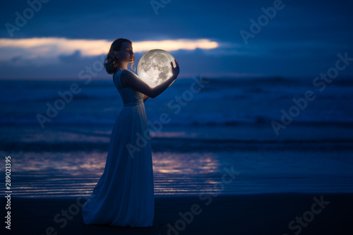 Tender image of a girl. Female magic. Beautiful attractive girl in full growth on a night beach with sand hugs the moon, art photo. On a dark background with space. Riddle, astrology, zodiac. Banner. photo