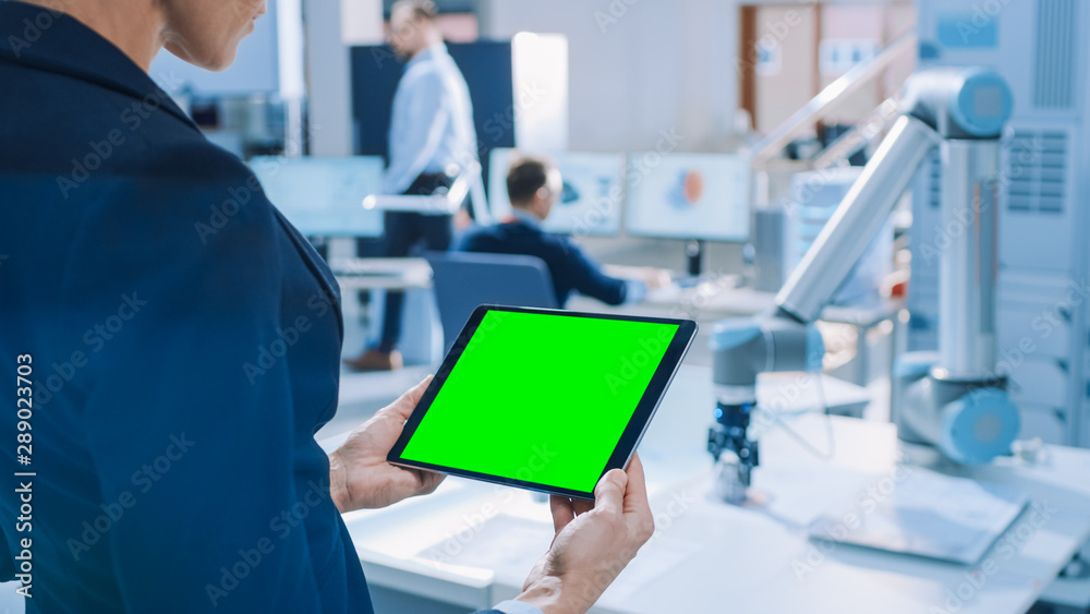 Engineer Holds Green Mock-up Screen Digital Tablet Computer. In the Background Team of Industrial Engineers Working with Functional Robot Arm in Modern Bright Research Facility Engineer Holds Green Mock-up Screen Digital Tablet Computer. In the Background Team of Industrial Engineers Working with Functional Robot Arm in Modern Bright Research Facility