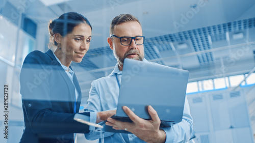 Stylish Confident Businesswoman and Businessman Working on a Project, Man Holds Laptop Computer, They Reference it. In the Background Modern and Bright Facility. Low Angle Shot photo