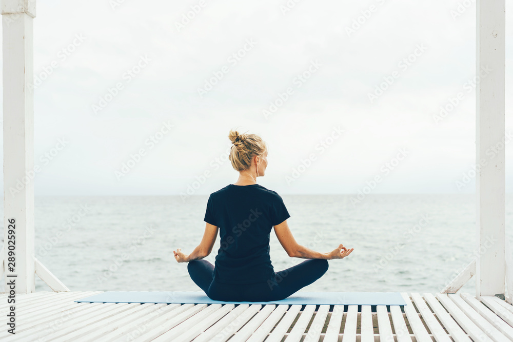A woman in sportswear is sitting in a lotus position with her back to the camera and meditating while looking at the sea. Calm peaceful mood. Mental comfort.