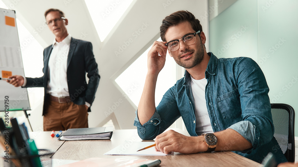Smart and confident. Young and handsome man adjusting eyeglasses and looking at camera with smile while his boss standing near whiteboard and explaining something on the background