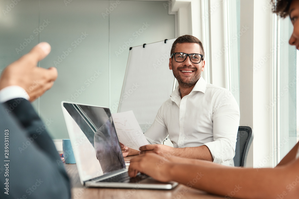 Fototapeta premium We have great results Young man in formal wear and eyeglasses holding financial report and smiling while sitting with his colleagues at the office table