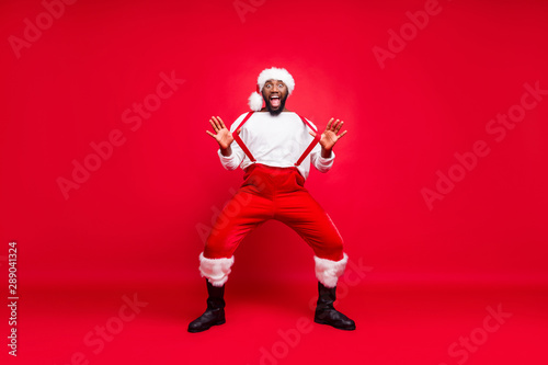 Full body photo of cheerful hipster afro american santa touching his suspenders screaming wearing trousers pants white pullover isolated over red background