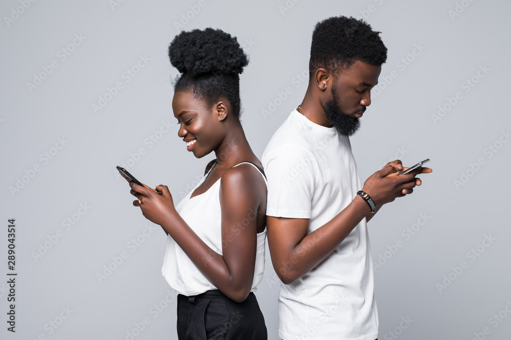 Happy young african american couple back to back laughing using smartphones apps isolated on gray studio background Happy young african american couple back to back laughing using smartphones apps isolated on gray studio background