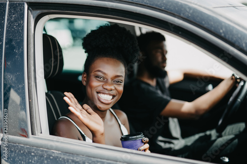 Smiling african couple in car with coffee. Young african girl looking at camera and greeting someone while drink coffee. African man at the wheel drive car