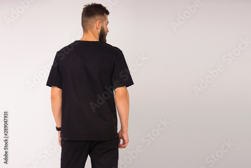 young stylish nurse male in black medical costume is standing from the back and looking away on white wall background. medical fashion concept. free space on right side