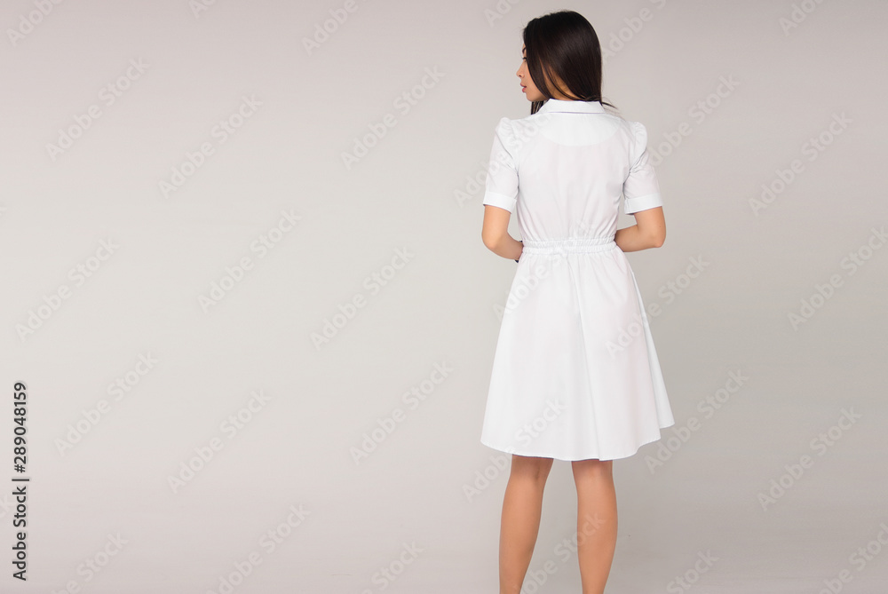 young stylish brunette nurse in white medical dress is standing from the back and looking away on the gray background. medical fashion concept, free space on left side young stylish brunette nurse in white medical dress is standing from the back and looking away on the gray background. medical fashion concept, free space on left side
