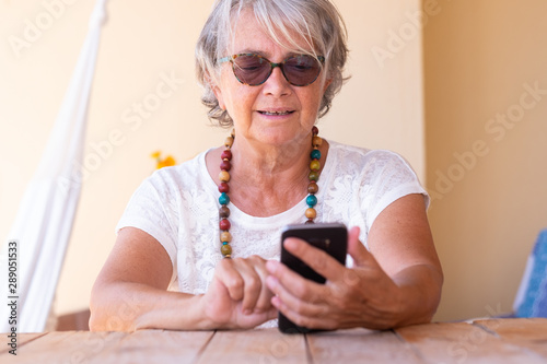Attractive senior woman messaging with telephone and smiling. Grey hair and sunglasses. Serene people sitting in the terrace. Wooden table