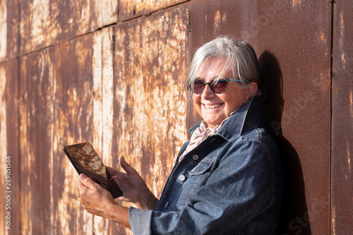 One smiling female people, senior woman with gray hair, holding the tablet and looking at camera. leaning against a rusted iron panel. Sunset light. Jeans jacket for the first cold days of autumn