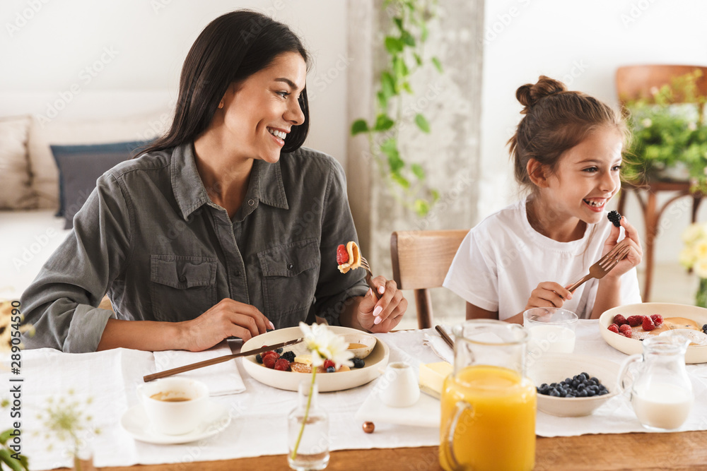 Image of joyous family mother and little daughter eating tasty food together while having breakfast at home in morning Image of joyous family mother and little daughter eating tasty food together while having breakfast at home in morning