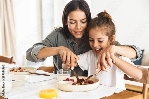 Image of amazing family mother and little daughter eating together while having breakfast at home in morning