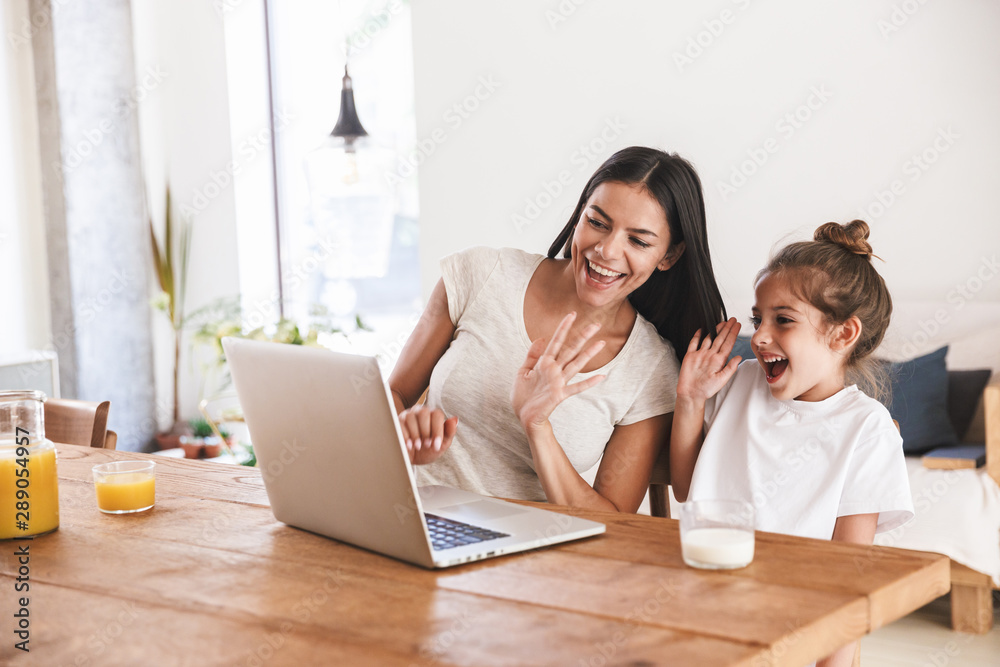 Image of caucasian family woman and her little daughter smiling and waving at laptop computer together in apartment Image of caucasian family woman and her little daughter smiling and waving at laptop computer together in apartment
