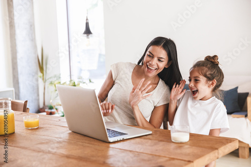 Image of caucasian family woman and her little daughter smiling and waving at laptop computer together in apartment
