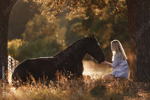 portrait of beautiful young woman with blond hair sitting in front of lying horse and feed it from hands in sunset sunlight in autumn