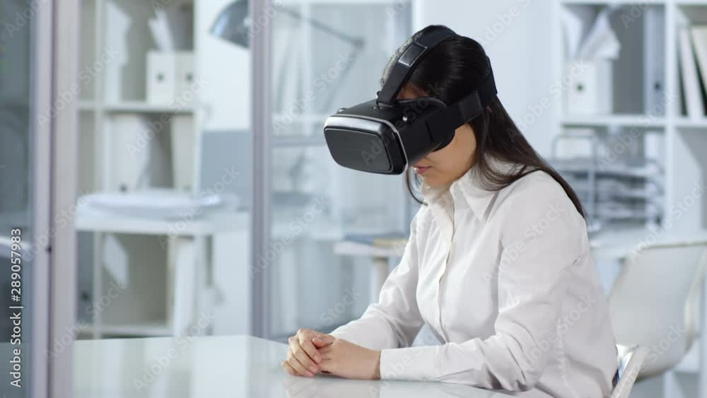 Waist-up shot of Asian woman with virtual reality headset sitting at desk in office, tapping on invisible interactive screen, looking at simulation and moving head around. Suitable for adding AR, VR. Waist-up shot of Asian woman with virtual reality headset sitting at desk in office, tapping on invisible interactive screen, looking at simulation and moving head around. Suitable for adding AR, VR.