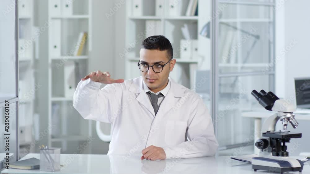 Waist-up shot of Middle Eastern male researcher in white lab coat sitting at desk with microscope beside, projecting invisible dimensional AR object and studying it attentively. Suitable for AR, VR. Waist-up shot of Middle Eastern male researcher in white lab coat sitting at desk with microscope beside, projecting invisible dimensional AR object and studying it attentively. Suitable for AR, VR.