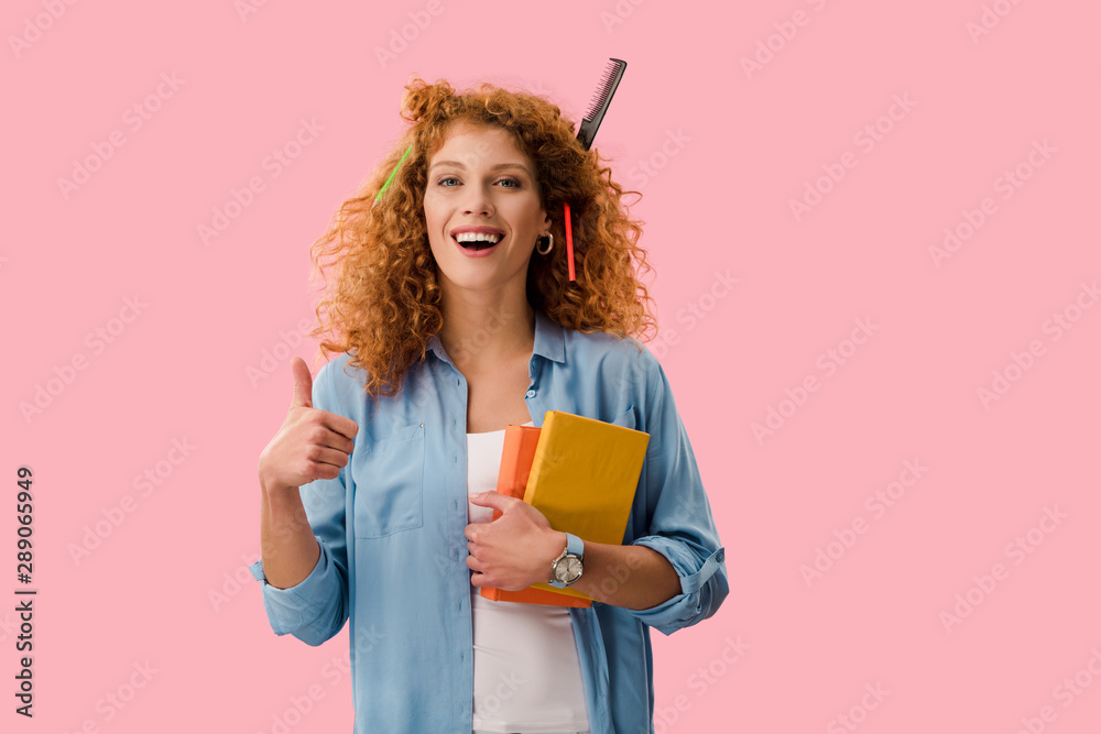 redhead student with pencils in hair holding books and showing thumb up isolated on pink redhead student with pencils in hair holding books and showing thumb up isolated on pink