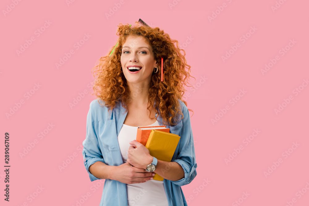 happy redhead student with pencils in hair holding books isolated on pink happy redhead student with pencils in hair holding books isolated on pink