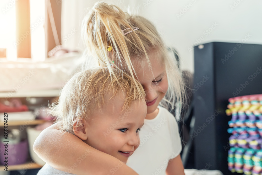 Cute adorable blond caucaian little sister hugging her toddler brother.couple of cheerful sibling playing indoor. Cute girl and baby boy enjoy relaxing at home. Happy childhood, family love, and care