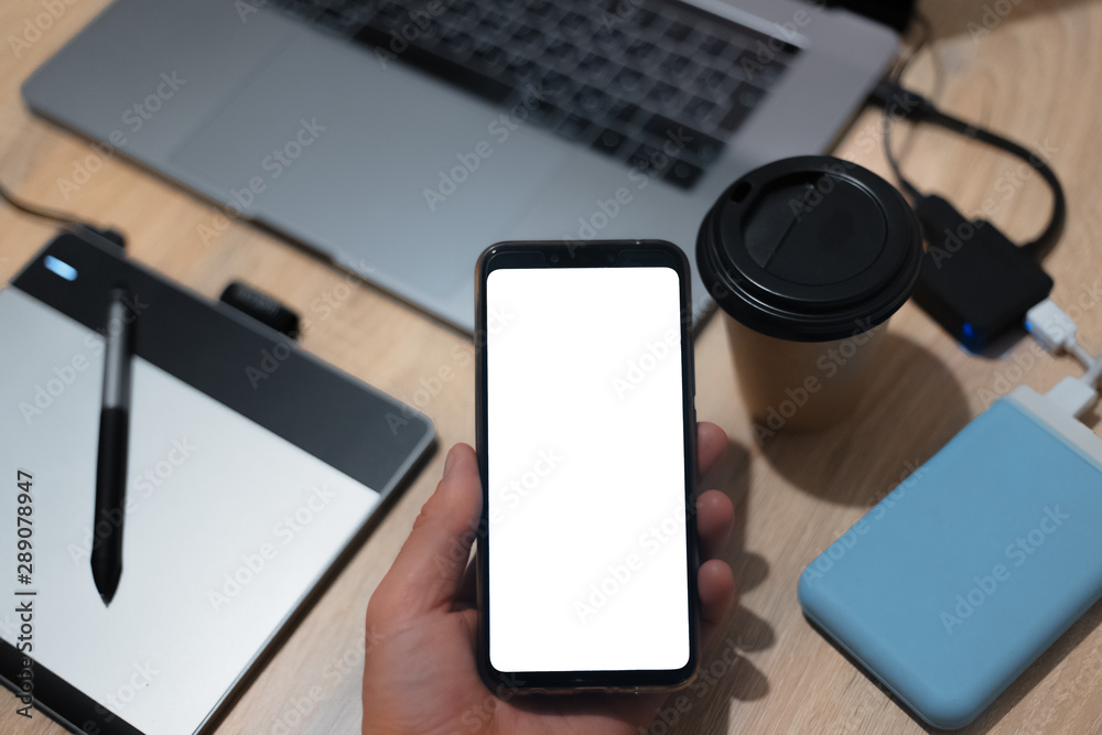 Male hand holding a smartphone with mockup. On background of designer desk with laptop, graphic tablet, cup of coffee, hard drive disk and type C adapter. Male hand holding a smartphone with mockup. On background of designer desk with laptop, graphic tablet, cup of coffee, hard drive disk and type C adapter.