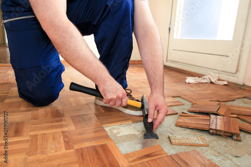 Construction worker and constructor is working on renovation of apartment. Handyman is removing old wooden parquet flooring using yellow hammer and scraping tool. Builder dismantles parquet floor.