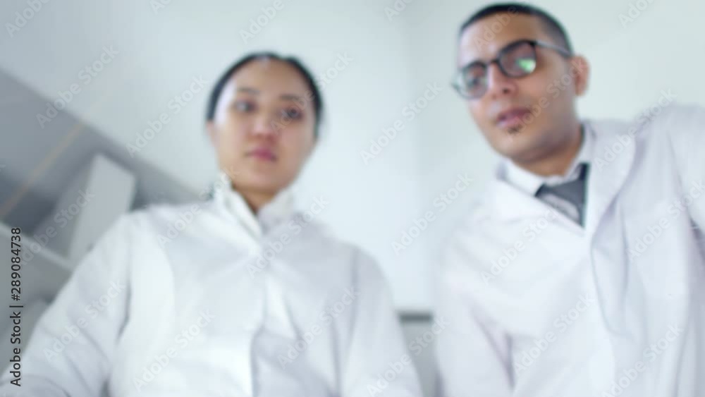 Low angle medium shot of Asian female and Arab male scientists in white lab coats standing above transparent AR touchscreen and looking at it with sweeping and tapping gestures. Suitable for AR, VR. Low angle medium shot of Asian female and Arab male scientists in white lab coats standing above transparent AR touchscreen and looking at it with sweeping and tapping gestures. Suitable for AR, VR.