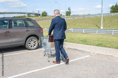 Elderly single man in a blue suit walks to his car with a supermarket trolley. Businessman and shopping