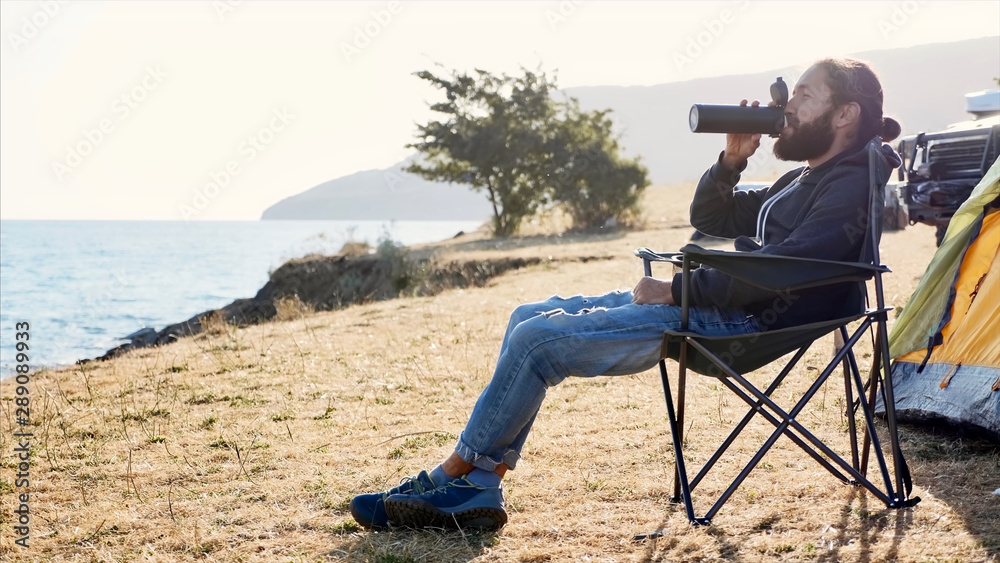 Young bearded hipster man sits on folding chair at campsite on lake shore and drinking tea or coffee from thermos bottle at morning. Side view with backlit