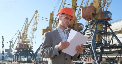 Young male worker of sea harbor in helmet, cargo manager in suit and halmet works outdoor , cranes and sea background	 photo