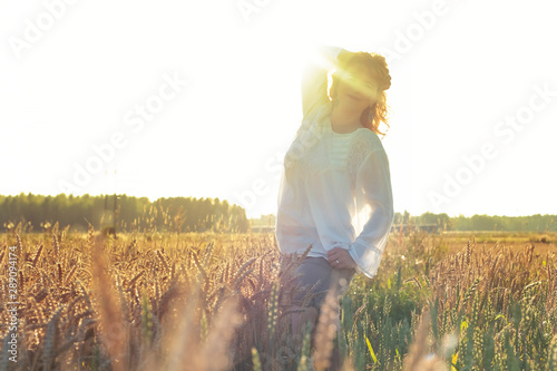   A girl with wavy red hair is standing in a field with wheat. Rural landscape. Blonde european girl watching sunset