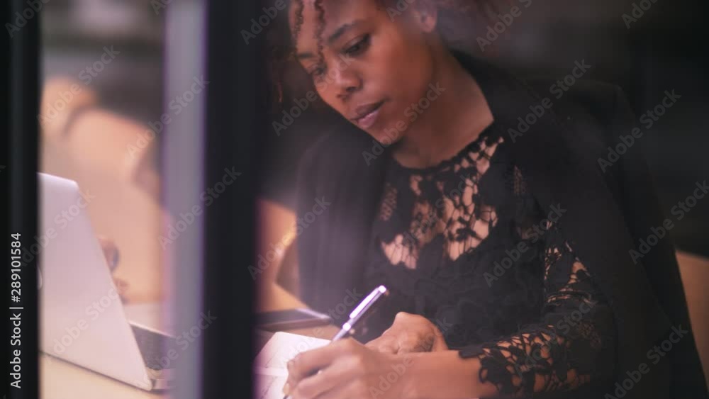 Through the window view of one young African female freelance writer writing in the cafe by the window young black businesswoman study learning working in urban office