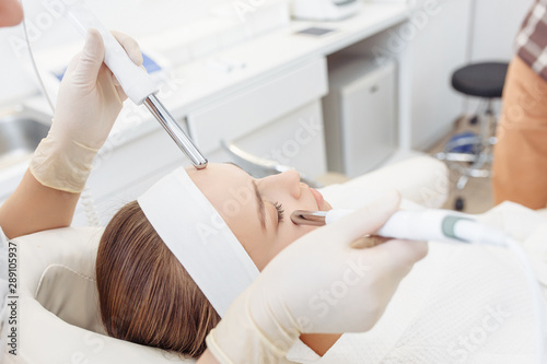 Beautician makes a microcurrent massage of the face of the girl client with the help of modern salon equipment. The concept of hardware cosmetology