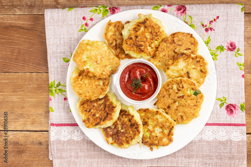Fritters lie on a white plate. In the middle is a gravy boat with tomato sauce. A dish stands on a wooden table covered by a napkin. Fritters made from zucchini.