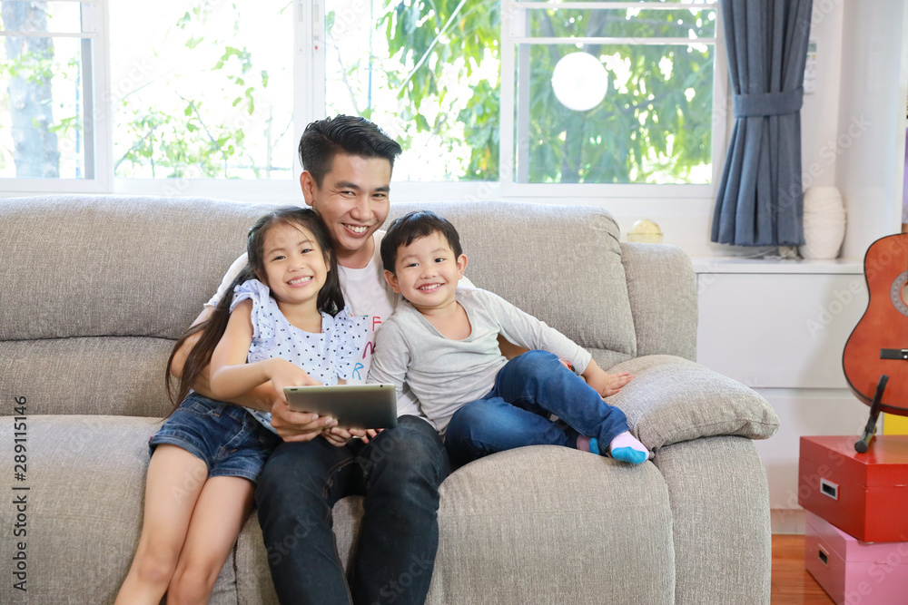 happy asian single dad teaching his daughter and son to use or play tablet on grey sofa in living room with laughing and smiling face (lifestyle with technology concept) happy asian single dad teaching his daughter and son to use or play tablet on grey sofa in living room with laughing and smiling face (lifestyle with technology concept)