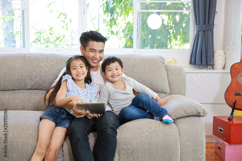 happy asian single dad teaching his daughter and son to use or play tablet on grey sofa in living room with laughing and smiling face (lifestyle with technology concept)