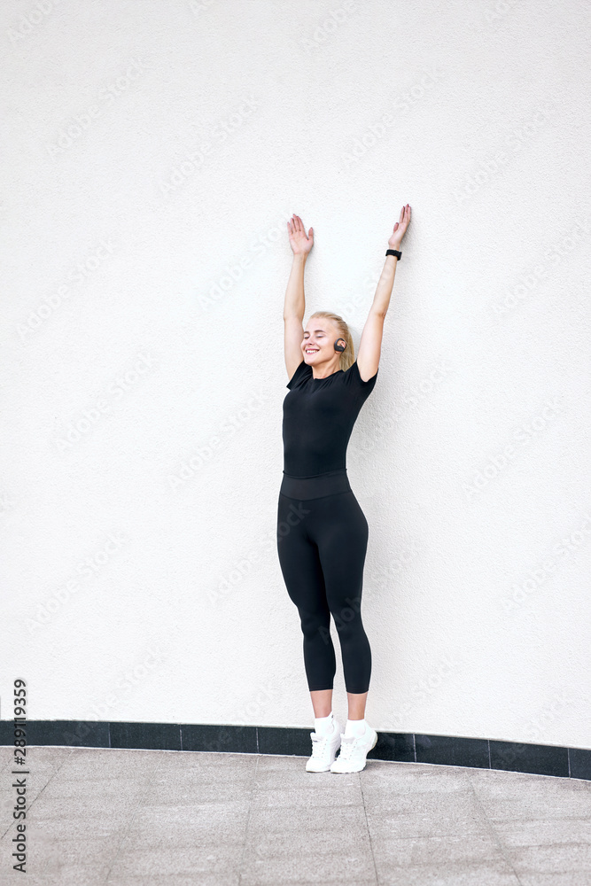 Young attractive sporty girl wearing black sportswear listening to music and stretching on white wall background. Concept active and healthy life.