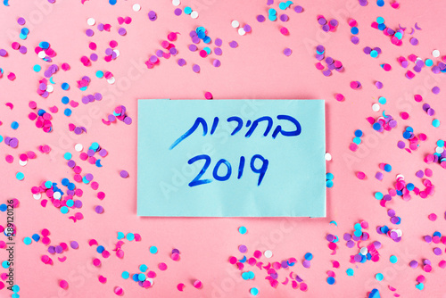 Hebrew text Elections 2019 on voting paper over pink with colorful confetti background. Israeli legislative Elections for the 21st Knesset Israel 2019.