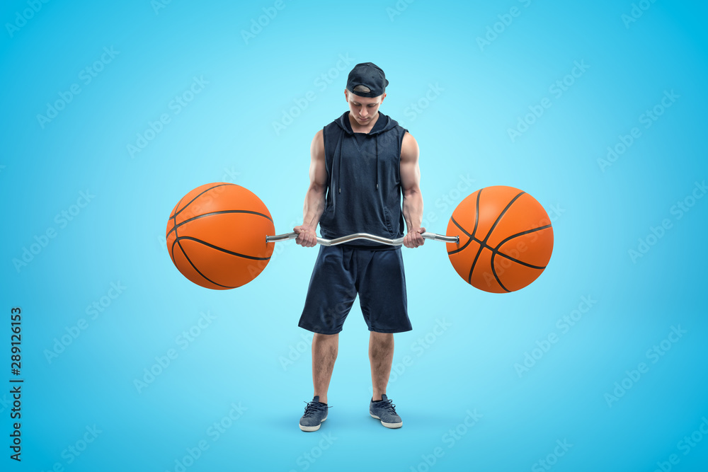 Front view of young athlete in black cap, sleeveless hoodie and shorts, standing and looking down at barbell in his hands, with two basketballs instead of weight plates.
