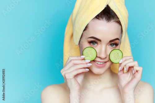 Portrait of young beautiful woman with healthy glow perfect smooth skin holds two piece of cucumber near face. Model with natural nude make up, with towel on head.