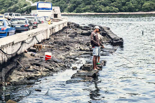 Phoograph of two poor boys fishing with a bamboo fishing rod, in a lagoon near Havana. In the background the flowing traffic. Vintage pastel colors