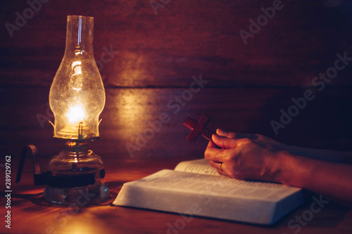 Close up of woman hands hold wooden cross over open bible with oil lamp on wood table while reading bible in the dark room, Christian background devotion or bible study concept