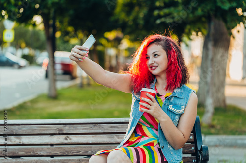 Stylish happy young woman in a bright striped sundress and a denim T-shirt with red hair sits on a bench with a phone and takes a selfie. sunny day